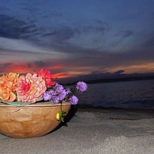 Pu&ograve; includere: Una ciotola di legno piena di una variet&agrave; di fiori colorati, tra cui arancio, rosa e viola, sullo sfondo di un tramonto su uno specchio d'acqua.