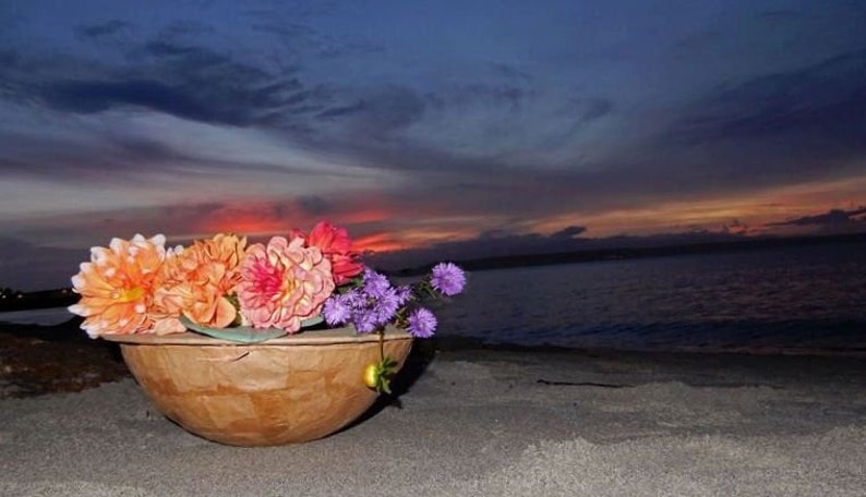Pu&ograve; includere: Una ciotola di legno piena di una variet&agrave; di fiori colorati, tra cui arancio, rosa e viola, sullo sfondo di un tramonto su uno specchio d'acqua.