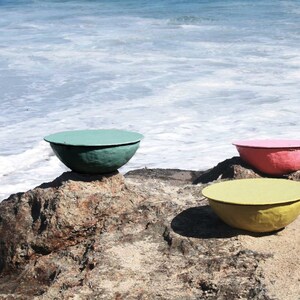 Puede incluir: Tres cuencos de cer&aacute;mica de colores, uno azul verdoso, uno amarillo y uno rosa, est&aacute;n sentados en una playa rocosa cerca del oc&eacute;ano.