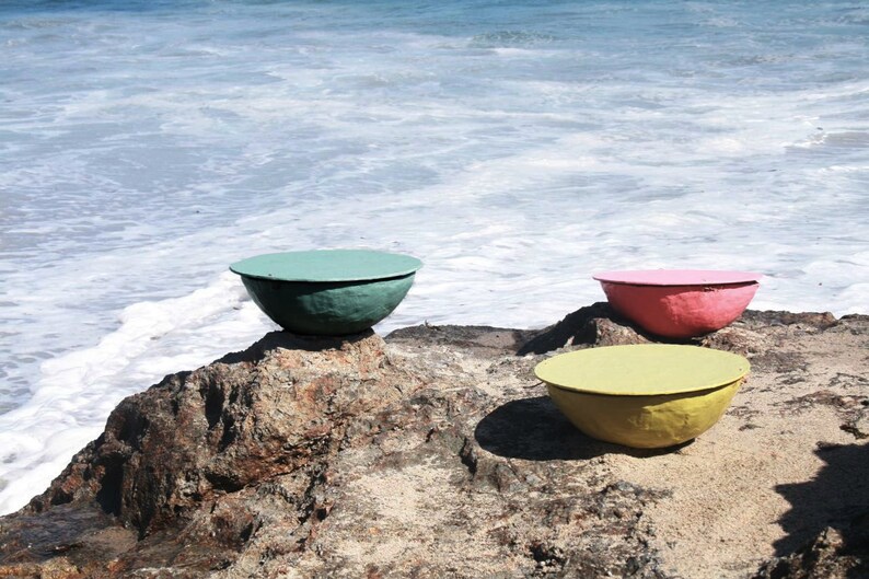 Puede incluir: Tres cuencos de cer&aacute;mica de colores, uno azul verdoso, uno amarillo y uno rosa, est&aacute;n sentados en una playa rocosa cerca del oc&eacute;ano.