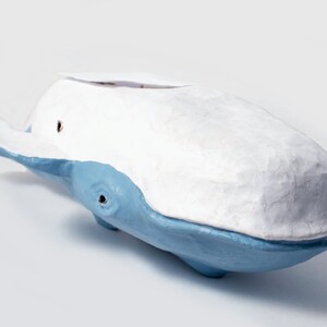 May include: A white and blue ceramic whale sculpture. The whale is painted in a light blue color with a white top. The whale has a small hole in the top of its head.