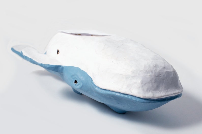 May include: A white and blue ceramic whale sculpture. The whale is painted in a light blue color with a white top. The whale has a small hole in the top of its head.