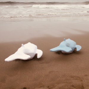 May include: Two ceramic whale sculptures on a sandy beach. One whale is white and the other is light blue.