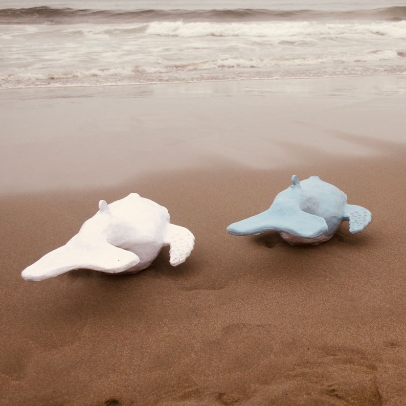 May include: Two ceramic whale sculptures on a sandy beach. One whale is white and the other is light blue.