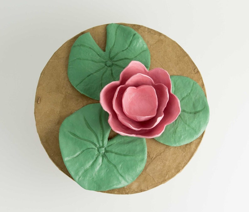 Pode incluir: Uma flor de l&iacute;rio d'&aacute;gua rosa com tr&ecirc;s folhas de l&iacute;rio d'&aacute;gua verdes sobre um fundo marrom.