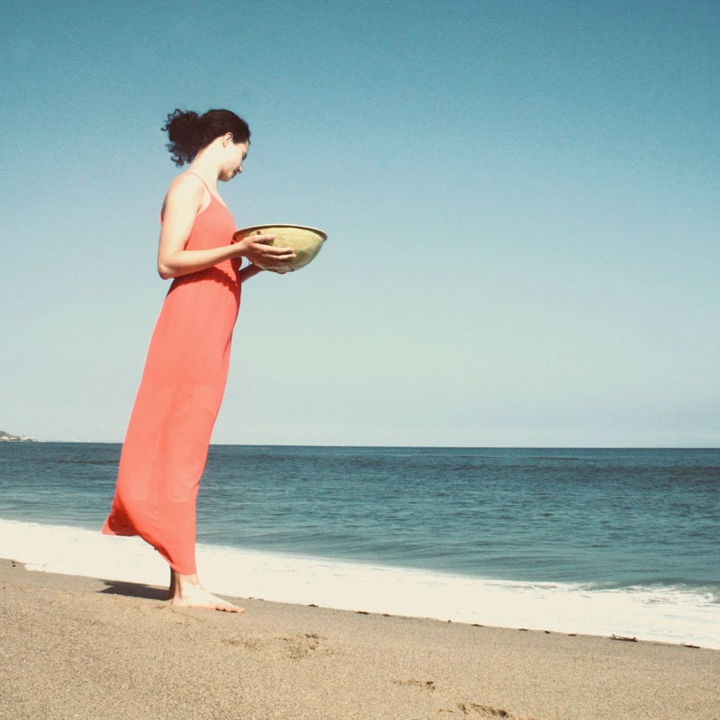 Puede incluir: Una mujer con un vestido rojo se encuentra en una playa de arena, sosteniendo un cuenco de cer&aacute;mica amarillo. El oc&eacute;ano est&aacute; en el fondo.