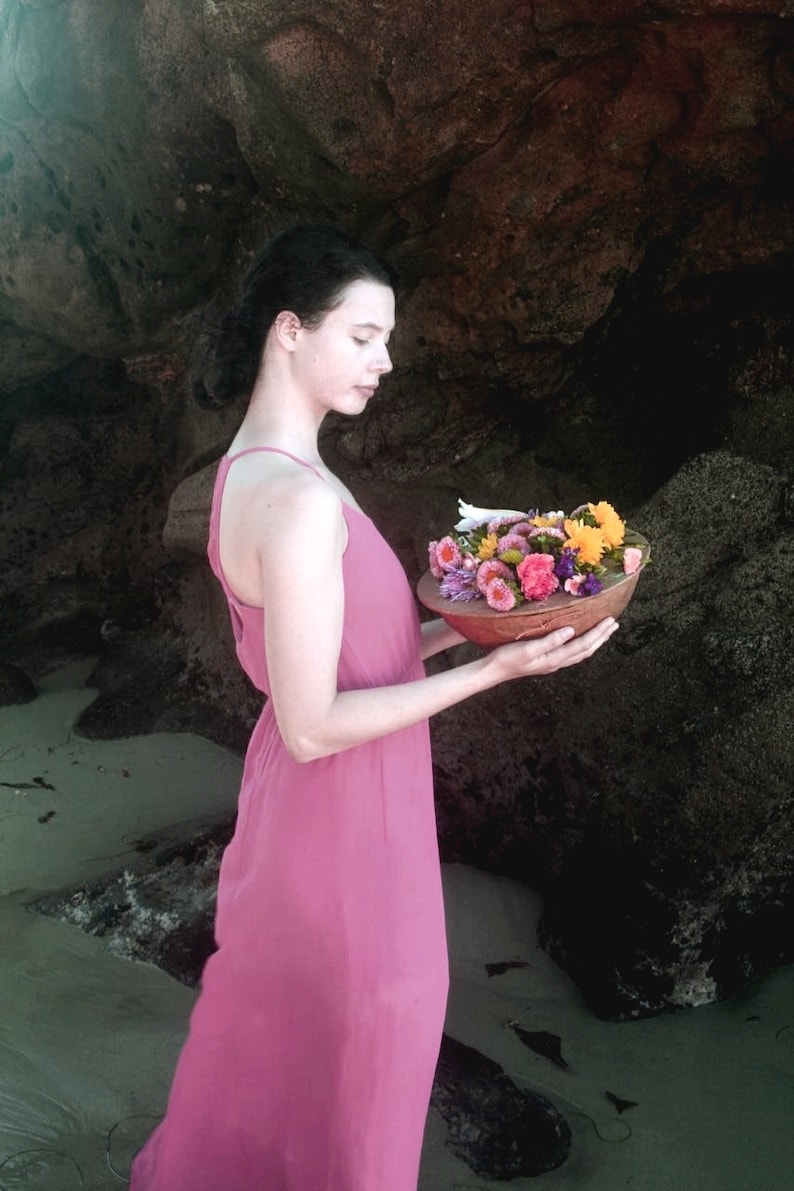 Pu&ograve; includere: Una donna con un vestito rosa tiene una ciotola di fiori colorati. La ciotola &egrave; in legno e ha una finitura naturale. La donna &egrave; in piedi davanti a una grande formazione rocciosa.