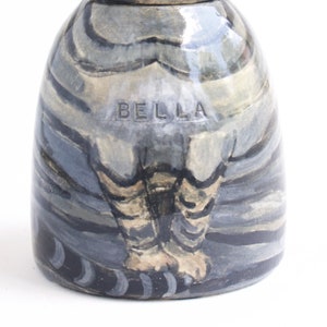 May include: A ceramic figurine of a woman with a blue and grey striped dress and black and white striped legs. The figurine is signed "BELLA" on the front.