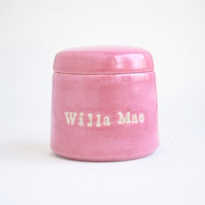 May include: A pink ceramic jar with a lid, featuring the name "Willa Mae" inscribed in white lettering.