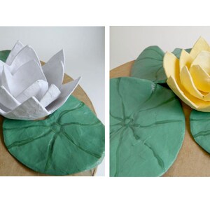 Pode incluir: Duas flores de l&iacute;rio d'&aacute;gua de papel mach&ecirc;, uma branca e outra amarela, est&atilde;o sentadas em cima de duas folhas de l&iacute;rio d'&aacute;gua de papel mach&ecirc; verdes.