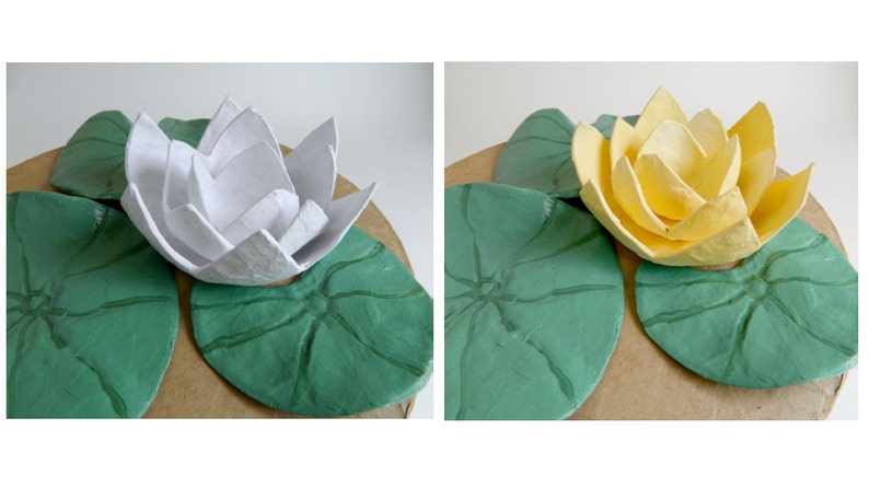 Pode incluir: Duas flores de l&iacute;rio d'&aacute;gua de papel mach&ecirc;, uma branca e outra amarela, est&atilde;o sentadas em cima de duas folhas de l&iacute;rio d'&aacute;gua de papel mach&ecirc; verdes.