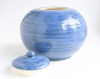 Op de afbeelding: Een blauwe keramische pot met een deksel. De pot is rond en heeft een gladde, geglazuurde afwerking. Het deksel is ook rond en heeft een knop bovenop.