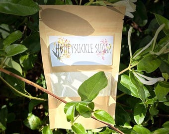 May include: A tan, rectangular, resealable pouch with a clear window at the bottom. A white label is centered on the upper portion of the pouch, featuring the text HONEYSUKKLE SUGAR in black, accompanied by a small floral illustration.