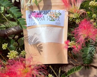 May include: A tan pouch containing white sugar, labeled HONEYSUCKLE & MIMOSA SUGAR, positioned among pink mimosa flowers and green leaves. The label includes the text NET WT. 4OZ and ORGANIC. The pouch has a clear window showing the white contents.