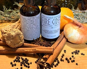 May include: Two amber glass bottles of Appalachian Homestead Fire Cider with black caps. Labels feature text including ingredients and instructions. Surrounded by raw ginger root, cinnamon sticks, star anise, black peppercorns, an orange, and an onion.