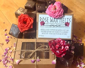 May include: A collection of dark brown wax melt cubes accompanied by a cardboard box with a window, tied with twine. The label reads ROSE WAX MELTS SOFT SWEET ROMANTIC, listing ingredients and warnings. Three fresh roses and scattered petals surround the items.