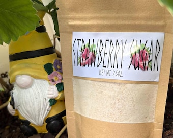 May include: A brown paper pouch containing strawberry sugar with a white label that reads STRAWBERRY SUGAR and NET WT. 2.5OZ. The label features a graphic of two strawberries. The pouch is positioned next to a yellow gnome figurine.