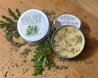 May include: Two round silver metal tins containing Bug Off Botanical Salve. One tin is closed with a white label featuring green herbs and purple flowers. The other tin is open, revealing a pale yellow balm infused with dried green herbal particles.