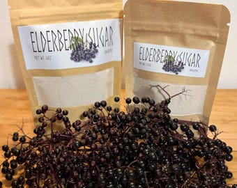 May include: Two brown paper pouches labeled Elderberry Sugar with white labels and clear windows, containing a light-colored powder. The pouches are positioned behind a large cluster of fresh, dark purple elderberries on thin stems.