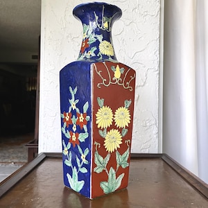 May include: A tall, rectangular ceramic vase with a flared top. The vase is divided into blue and red panels, each decorated with floral patterns in yellow, red, and green. The vase sits on a dark wooden surface.
