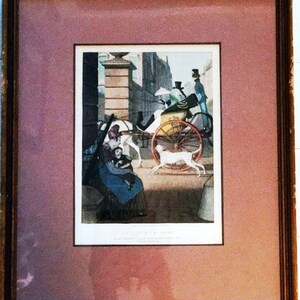 Op de afbeelding: Een ingelijste zwart-witprint van een door een paard getrokken koets met een vrouw en een kind op de voorgrond. De koets wordt bestuurd door een man met een hoge hoed. De scène speelt zich af in een straat in de stad met gebouwen op de achtergrond.