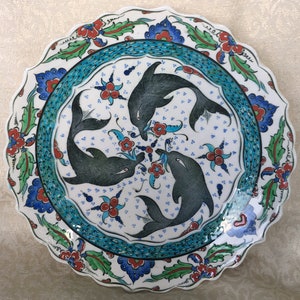 Puede incluir: Un plato de cerámica blanco con un borde festoneado presenta un diseño floral azul y verde alrededor del borde. El centro del plato representa tres delfines negros nadando en círculo, rodeados de flores rojas y azules.