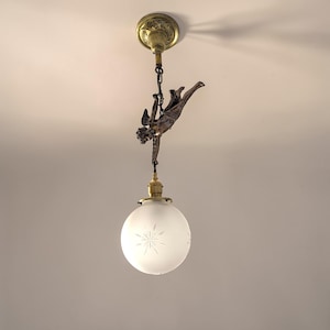 Peut inclure: Un luminaire de plafond de style ancien. Une figurine de chérubin en bronze est suspendue à un support de plafond en laiton. La lumière a un globe en verre dépoli avec un motif en forme d'éclat d'étoile.