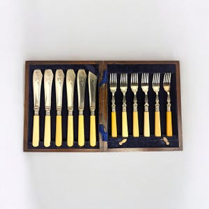 May include: A vintage cutlery set in a wooden box. The set includes knives and forks with cream-colored handles and ornate silver blades and tines. The box is lined with dark blue fabric.