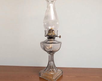 May include: A clear glass oil lamp with a tall, curved chimney and a decorative base. The lamp features a brass burner and a clear glass reservoir. The base is square with a gold-colored trim. The lamp is approximately 12 inches tall.