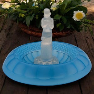 May include: A frosted glass figurine of a woman standing in a light blue glass bowl with a decorative pattern. The bowl is on a wooden surface.