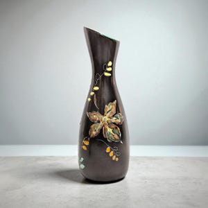 Puede incluir: Un jarrón de cerámica marrón oscuro con una parte superior asimétrica única. El jarrón presenta un diseño floral grande y detallado en tonos de marrón, amarillo y verde. Delicados acentos de hojas y puntos en amarillo y naranja adornan el jarrón.