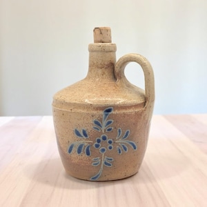 Peut inclure: Une cruche en céramique avec un bouchon en liège et un motif floral bleu. La cruche est de couleur brun clair avec un glaçage moucheté.