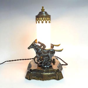 Puede incluir: Lámpara de mesa antigua en tono bronce con una figura de caballo y jinete. La lámpara tiene una pantalla cilíndrica de vidrio blanco, una parte superior de filigrana decorativa y una base detallada. La lámpara mide unos 30 cm de alto.