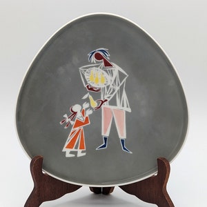 May include: A grey ceramic plate featuring a stylised illustration of two figures. The figure on the left is a child holding a pear, and the figure on the right is an adult holding a basket of pears. The plate is displayed on a wooden stand.
