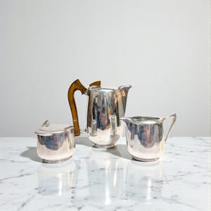 May include: A silver tea set comprising a teapot with a wooden handle, a creamer jug, and a lidded sugar bowl. The set is displayed on a white marble surface. The teapot's handle is made of wood.