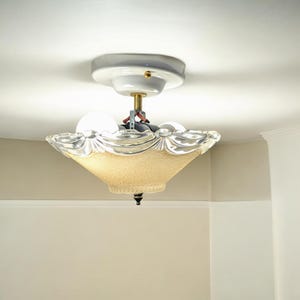 Pode incluir: Uma luminária de teto vintage com uma placa de teto branca e um abajur de vidro fosco com borda festonada. O abajur é de cor bege claro e tem um design decorativo.