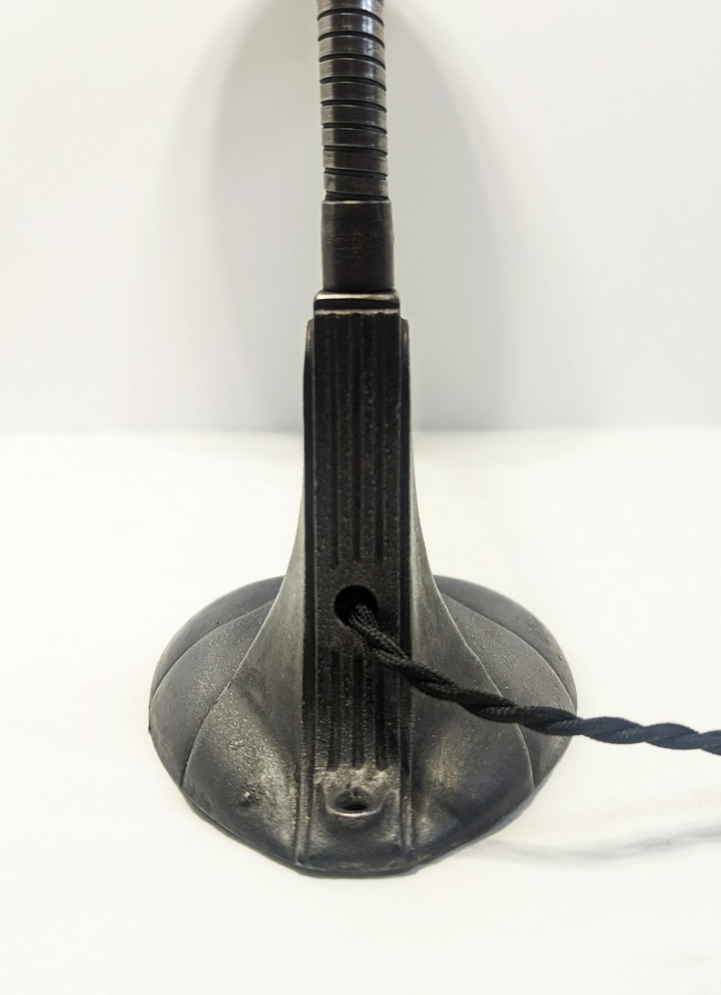 May include: Black metal desk lamp with a flexible arm and a round base. The lamp has a cord with a black braided wire.