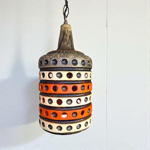 May include: A vintage pendant light fixture with a cylindrical shape. The lamp features alternating bands of orange, cream, and black, with circular cutouts. The top section is a textured brown and gray. The light hangs from a chain.