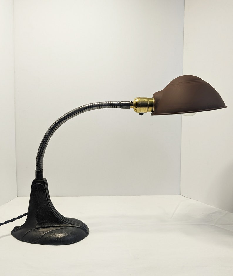 May include: A black metal desk lamp with a flexible gooseneck arm and a brown metal shade. The lamp has a classic design and is in good condition.