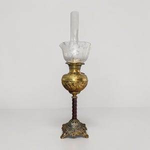 May include: An antique oil lamp with a clear glass chimney and a floral-etched glass shade. The lamp has a brass body with ornate detailing, a dark brown twisted stem, and a decorative base. A vintage home decor item.