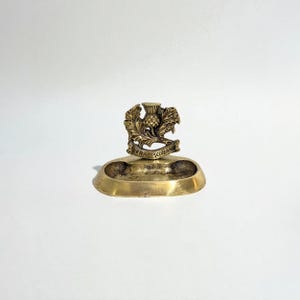 Puede incluir: Un cenicero de latón antiguo con un emblema de cardo. El emblema presenta un diseño detallado de cardo con las palabras "FRAE SCOTLAND" en una pancarta. El cenicero tiene forma ovalada y poco profunda.