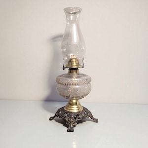May include: A vintage oil lamp with a clear glass chimney and reservoir. The reservoir has a decorative etched pattern. The lamp features a brass burner and a decorative metal base with three feet. The lamp is designed to sit on a flat surface.
