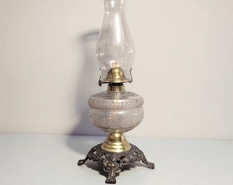 May include: A vintage oil lamp with a clear glass chimney and reservoir. The reservoir has a decorative etched pattern. The lamp features a brass burner and a decorative metal base with three feet. The lamp is designed to sit on a flat surface.