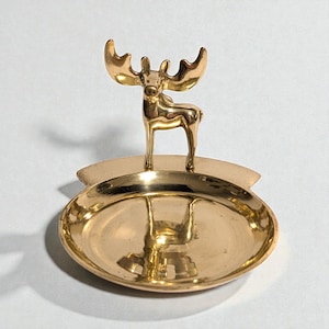 May include: A gold-coloured metal dish with a moose figurine. The dish is oval-shaped with a raised rim and a small moose figure standing in the centre. The moose has large antlers and is facing forward.