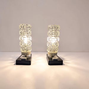Op de afbeelding: Een paar tafellampen met cilindrische, getextureerde helderglazen kappen met een geometrisch patroon. Elke lamp heeft een rechthoekige zwarte voet met een witte vierkante schakelaar en straalt warm licht uit vanuit het midden.