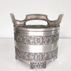 May include: A round silver-toned metal container with a lid and a central handle. The exterior features two horizontal bands of embossed floral and scrollwork patterns. The container is supported by three small, ornate feet and has two side handles.
