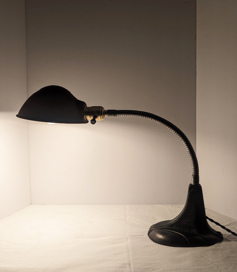 May include: A black metal desk lamp with a flexible arm and a dome-shaped shade. The lamp is on a white surface.
