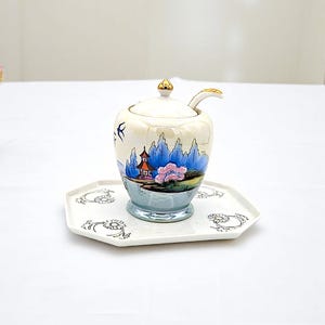 May include: A white porcelain sugar bowl with a lid and a matching tray. The bowl has a blue and gold floral design with a small red building in the center. The tray has a gold floral design.