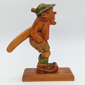 May include: A wooden nutcracker figurine of a man wearing a green hat and red shirt, holding a wooden lever in his hand. The figurine is standing on a wooden base.