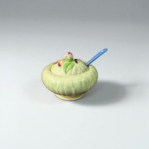 May include: A pale green ceramic sugar bowl with a lid, decorated with a floral motif and a blue spoon. The bowl has a textured, ribbed exterior and a small, round base. The lid features a pink flower bud and green leaves.